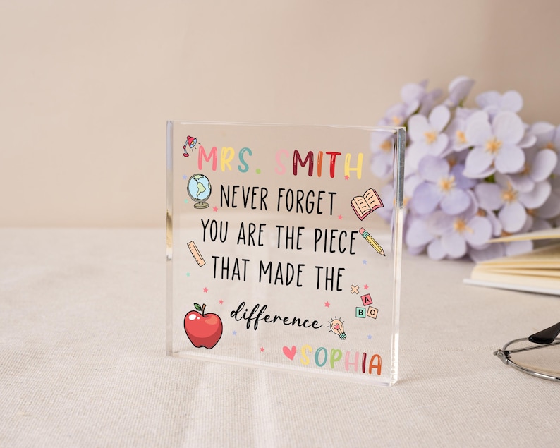 May include: A clear acrylic block with colorful text and illustrations. The text reads "MRS. SMITH NEVER FORGET YOU ARE THE PIECE THAT MADE THE difference SOPHIA." Includes a red apple, globe, and pencil. A thoughtful gift for a teacher.