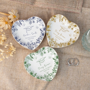 Könnte beinhalten: Drei herzförmige Keramikschalen mit Blumendesigns und Illustrationen von Abschlusskappen. Jede Schale hat einen goldenen Rand und personalisierten Text. Eine Schale ist blau, eine ist goldfarben und eine ist grün. Zwei silberne Ringe liegen neben den Schalen.