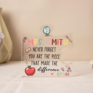 May include: Clear acrylic puzzle piece with the text "MRS. SMITH NEVER FORGET YOU ARE THE PIECE THAT MADE THE difference SOPHIA". The puzzle piece has a globe, pencil, apple, and book illustrations. The text and illustrations are in various colors.