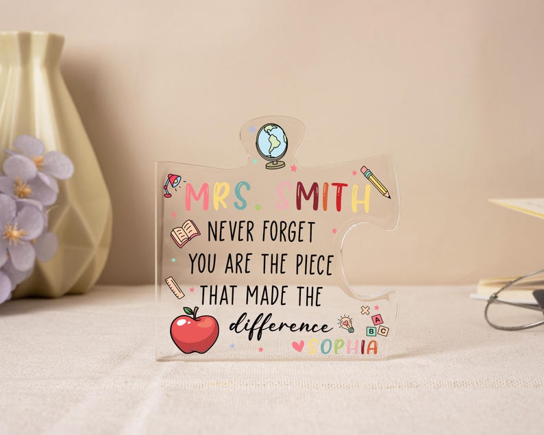 May include: Clear acrylic puzzle piece with the text "MRS. SMITH NEVER FORGET YOU ARE THE PIECE THAT MADE THE difference SOPHIA". The puzzle piece has a globe, pencil, apple, and book illustrations. The text and illustrations are in various colors.