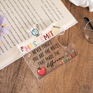 May include: A clear acrylic bookmark with the text "MRS. SMITH NEVER FORGET YOU ARE THE PIECE THAT MADE THE difference SOPHIA" and colorful illustrations. The bookmark is on a wooden surface next to a book, glasses, and flowers.