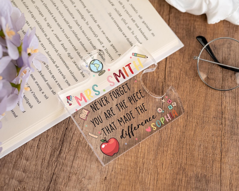 May include: A clear acrylic bookmark with the text "MRS. SMITH NEVER FORGET YOU ARE THE PIECE THAT MADE THE difference SOPHIA" and colorful illustrations. The bookmark is on a wooden surface next to a book, glasses, and flowers.