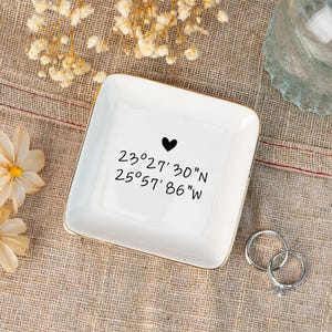 May include: A white square trinket dish with gold trim, featuring a black heart and coordinates. The coordinates are in black script. Two silver rings are next to the dish. The dish is on a tan surface.