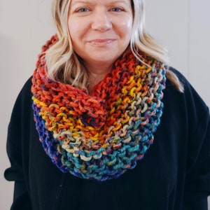 May include: A woman wearing a black sweater with a colourful crocheted infinity scarf. The scarf is made with a variety of colours, including red, orange, yellow, green, blue, and purple.