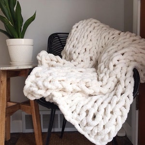 May include: A white chunky knit throw blanket draped over a black chair. The blanket is made of thick yarn and has a soft, fluffy texture.