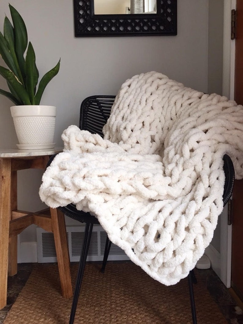 May include: A white chunky knit throw blanket draped over a black chair. The blanket is made of thick yarn and has a soft, fluffy texture.