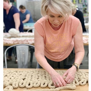 May include: A person is working on a large, beige, chunky knit blanket. The blanket is made with thick yarn and has a textured, looped design. The person is wearing a peach colored t-shirt with the text "You're doing great" printed on it.