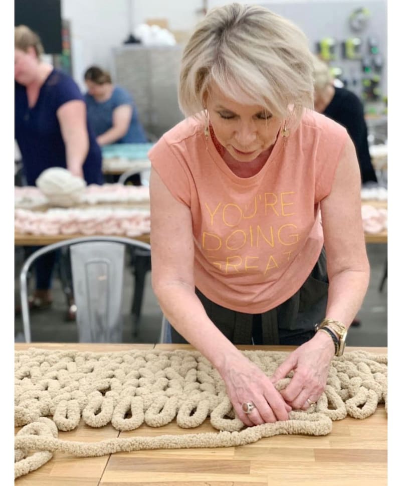 May include: A person is working on a large, beige, chunky knit blanket. The blanket is made with thick yarn and has a textured, looped design. The person is wearing a peach colored t-shirt with the text "You're doing great" printed on it.