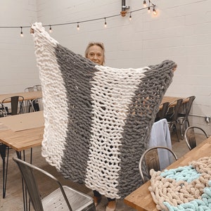 May include: A woman holds up a large, chunky knit blanket with alternating stripes of white and gray yarn. The blanket is made with thick, fluffy yarn and has a soft, cozy texture.