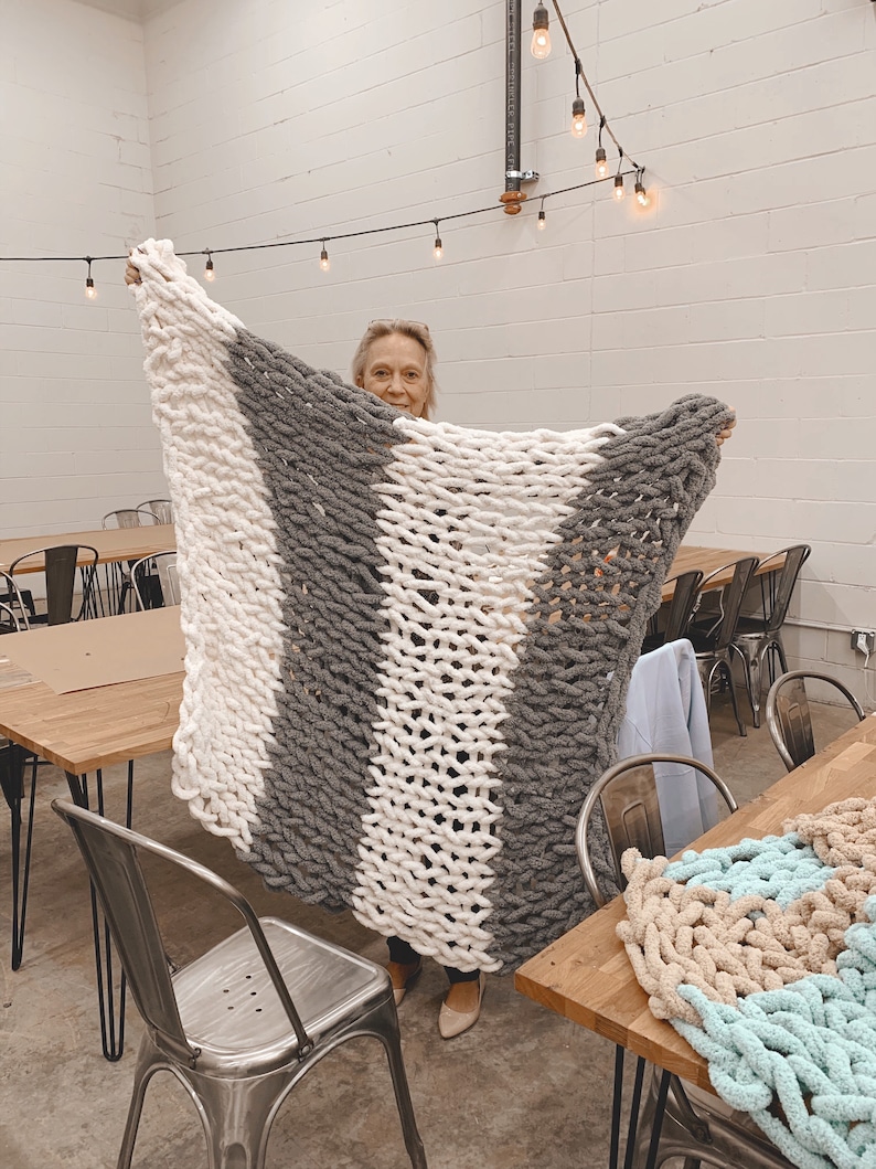 May include: A woman holds up a large, chunky knit blanket with alternating stripes of white and gray yarn. The blanket is made with thick, fluffy yarn and has a soft, cozy texture.