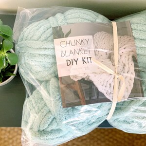 May include: A DIY chunky knit blanket kit in a clear plastic bag. The kit includes a large ball of light blue yarn and a smaller ball of white yarn. The label on the bag reads "Chunky Blanket DIY Kit by Kristin  Hall".