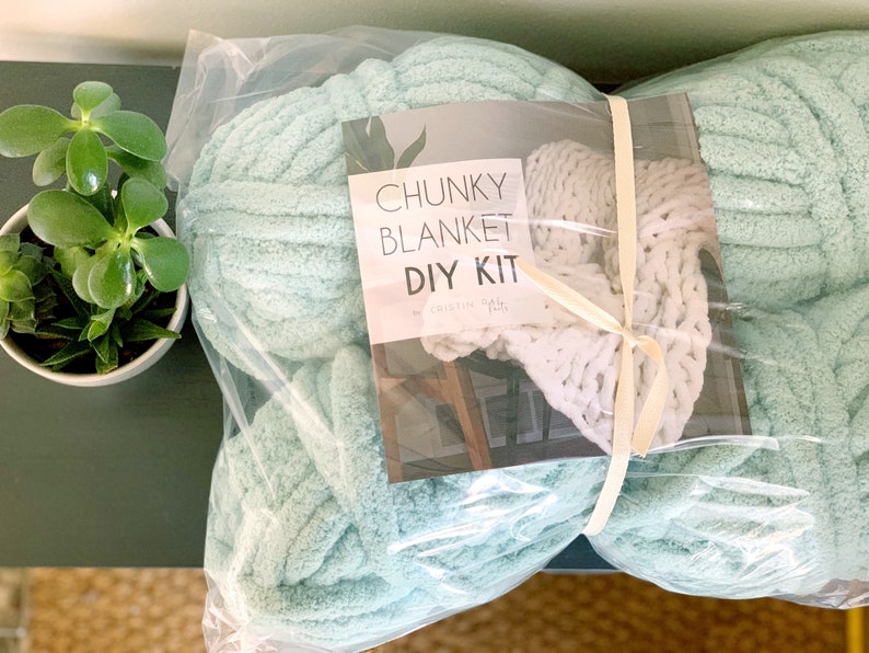 May include: A DIY chunky knit blanket kit in a clear plastic bag. The kit includes a large ball of light blue yarn and a smaller ball of white yarn. The label on the bag reads "Chunky Blanket DIY Kit by Kristin  Hall".