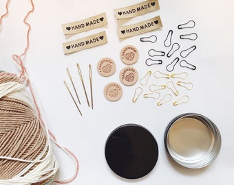 May include: A collection of craft supplies including yarn, needles, safety pins, and small leather tags that say "HAND MADE" in black lettering with a red heart.