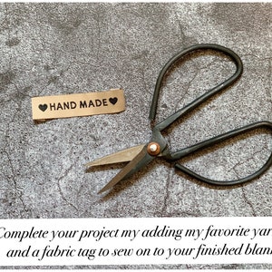 May include: A pair of black yarn scissors and a brown fabric tag with the text "HAND MADE" and a black heart on each side. The scissors and tag are on a gray textured surface.