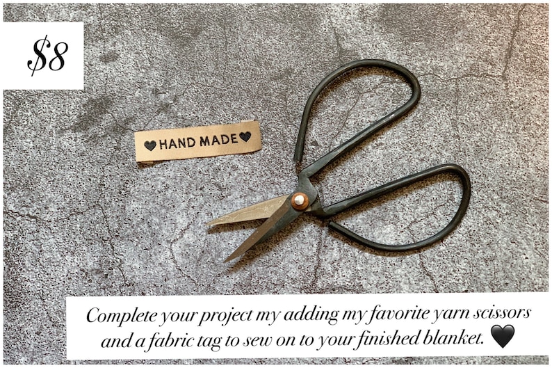 May include: A pair of black yarn scissors and a brown fabric tag with the text "HAND MADE" and a black heart on each side. The scissors and tag are on a gray textured surface.