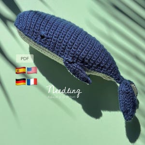 May include: A handmade, crocheted whale in shades of blue and gray. The whale is lying on a light green surface, with the word "Needling" and the phrase "Aguja y crochet" visible. The image also includes flags and the letters "PDF".