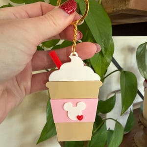 May include: A coffee-themed keychain with a gold ring and chain. The keychain features a tan coffee cup with a pink band, a white Mickey Mouse design, and a red heart. A red straw and white whipped cream top the cup.