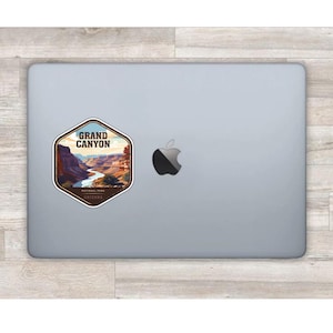 May include: A hexagonal sticker featuring a graphic illustration of the Grand Canyon with a winding river, layered rock formations, and the text "GRAND CANYON NATIONAL PARK ARIZONA" applied to the lid of a silver laptop with an Apple logo.