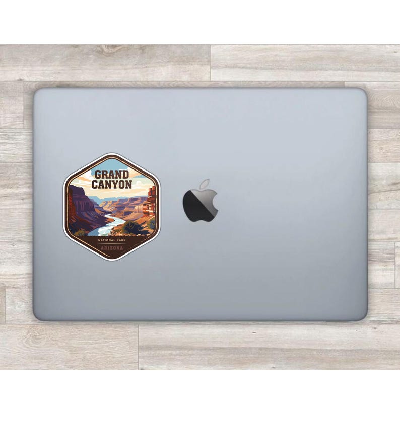 May include: A hexagonal sticker featuring a graphic illustration of the Grand Canyon with a winding river, layered rock formations, and the text "GRAND CANYON NATIONAL PARK ARIZONA" applied to the lid of a silver laptop with an Apple logo.