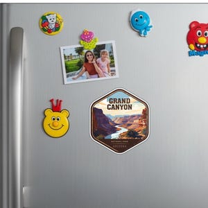 May include: A hexagonal sticker featuring a stylized illustration of the Grand Canyon in Arizona, with the text GRAND CANYON NATIONAL PARK ARIZONA. The sticker is displayed on a metallic surface alongside several colorful character magnets.