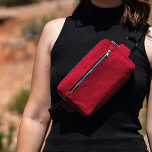 May include: A red fanny pack with a silver zipper is worn across the body. The bag is made of a textured fabric and has a black strap. The person is wearing a black tank top.