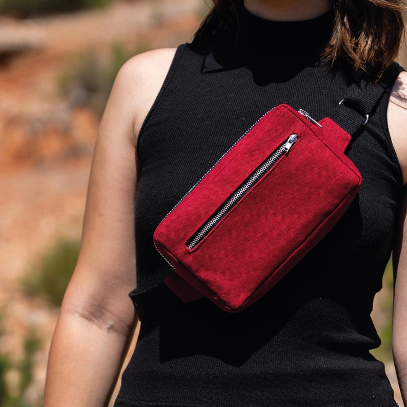 May include: A red fanny pack with a silver zipper is worn across the body. The bag is made of a textured fabric and has a black strap. The person is wearing a black tank top.