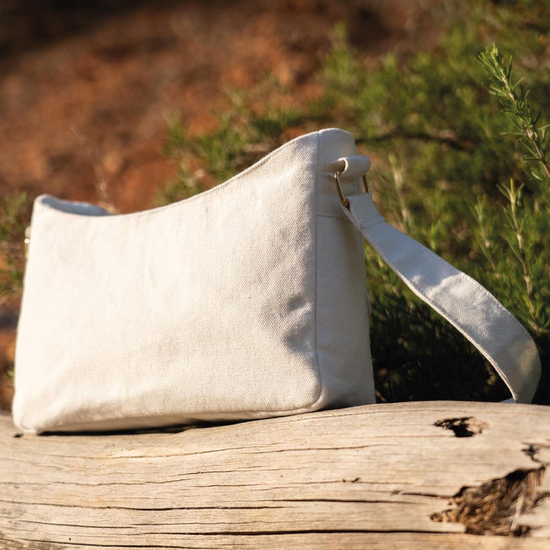 May include: A white canvas tote bag with a long strap. The bag is sitting on a wooden log.