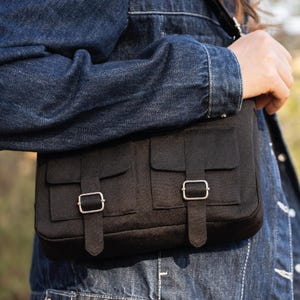 May include: A black canvas messenger bag with two front pockets and silver buckles. The bag is worn over the shoulder of a person wearing a denim jacket.