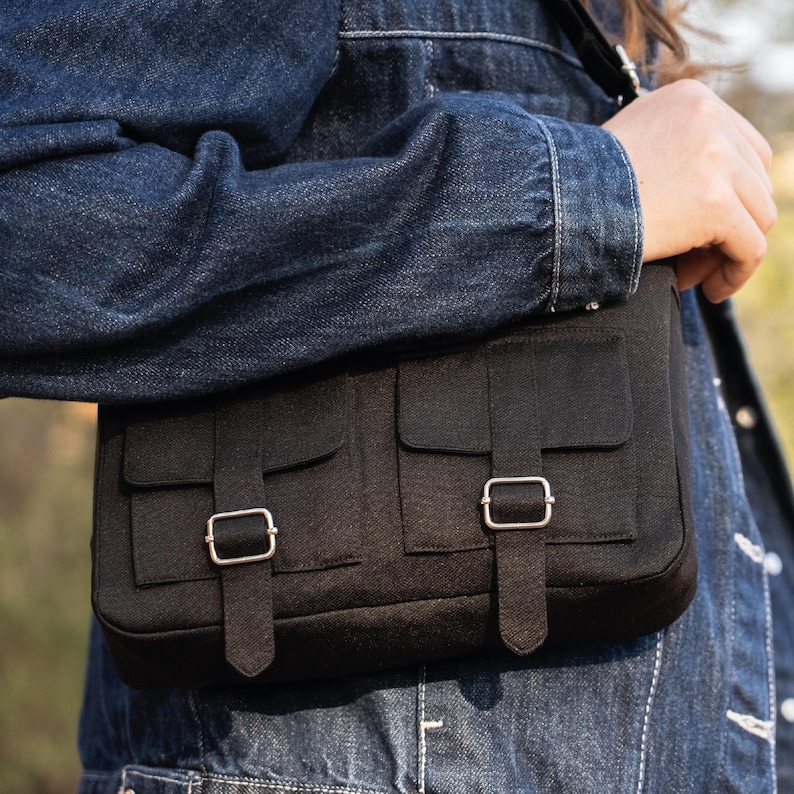 May include: A black canvas messenger bag with two front pockets and silver buckles. The bag is worn over the shoulder of a person wearing a denim jacket.