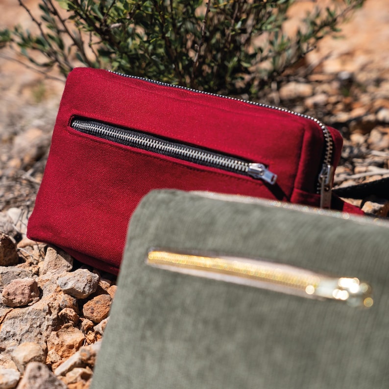 May include: A close-up of two rectangular zippered pouches. The top pouch is a deep red with a black zipper. The bottom pouch is a muted green with a gold zipper. Both pouches are made of a textured fabric and are set on a rocky surface.