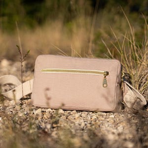 May include: A beige rectangular crossbody bag with a gold zipper and hardware. The bag has a matching beige strap and is set against a backdrop of dry grass and small rocks. The bag is made of a canvas-like material.