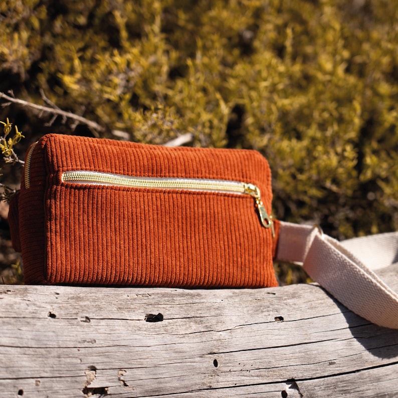 May include: A rust-colored corduroy fanny pack with a gold zipper and a cream-colored strap. The bag is rectangular and sits on a weathered log, with a blurred green background.