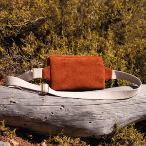 May include: A rust-colored corduroy fanny pack with a cream-colored adjustable strap and a silver-toned buckle. The bag is rectangular with a zippered top. The strap is made of a sturdy canvas material. The bag is resting on a weathered log.