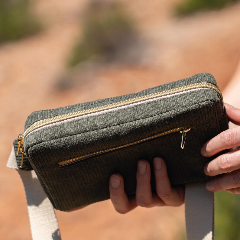 May include: A green corduroy fanny pack with a gold zipper and a white strap. The rectangular bag has a small front pocket and is held by a person. The background is blurred, suggesting an outdoor setting.