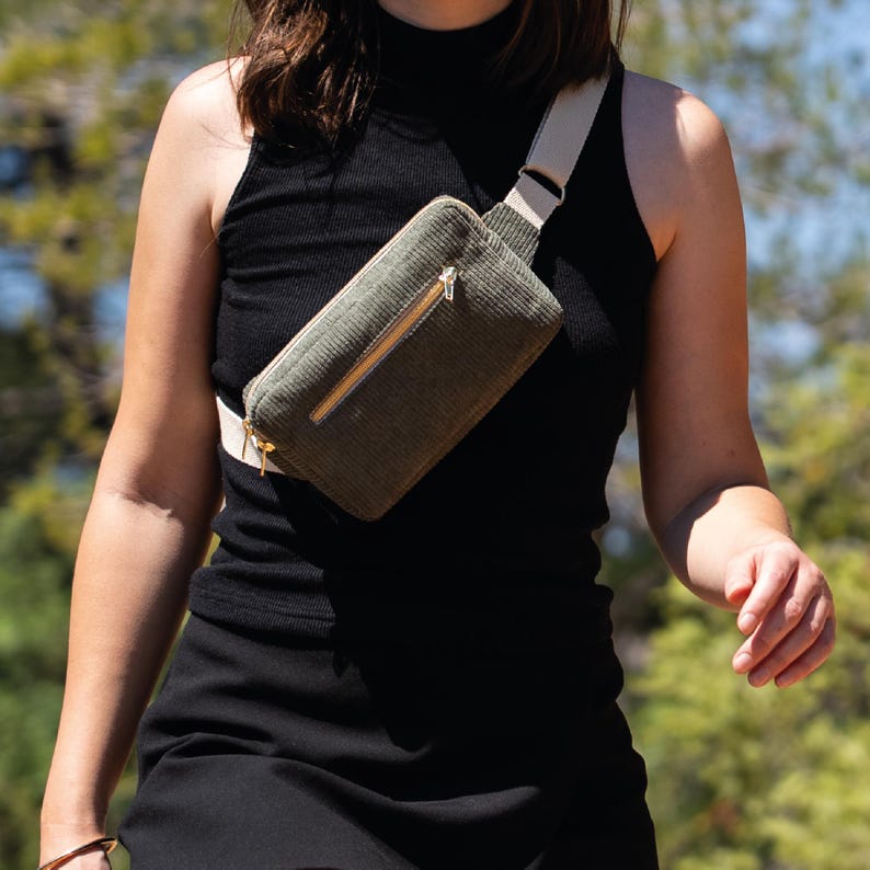 Sac banane en velours côtelé vert olive avec une sangle beige et une fermeture éclair dorée. Le sac est porté en bandoulière.