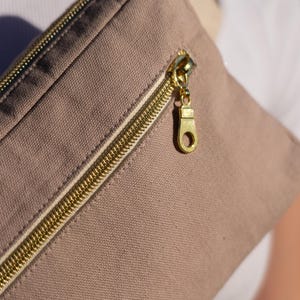 May include: Close-up of a tan canvas fanny pack with a gold zipper. The zipper pull is also gold. The fanny pack is made of a durable fabric and has a simple, minimalist design.
