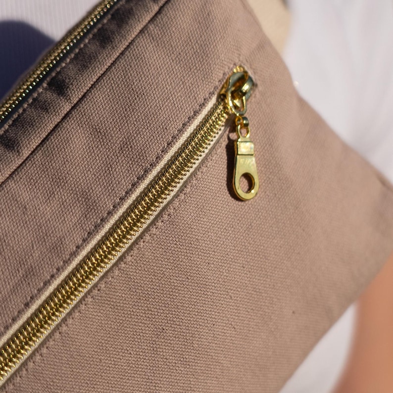 May include: Close-up of a tan canvas fanny pack with a gold zipper. The zipper pull is also gold. The fanny pack is made of a durable fabric and has a simple, minimalist design.