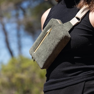 May include: Olive green corduroy fanny pack with a gold zipper. The bag has a beige strap and is worn across the body. The bag is rectangular and has a minimalist design. The fanny pack is a stylish accessory for carrying essentials.