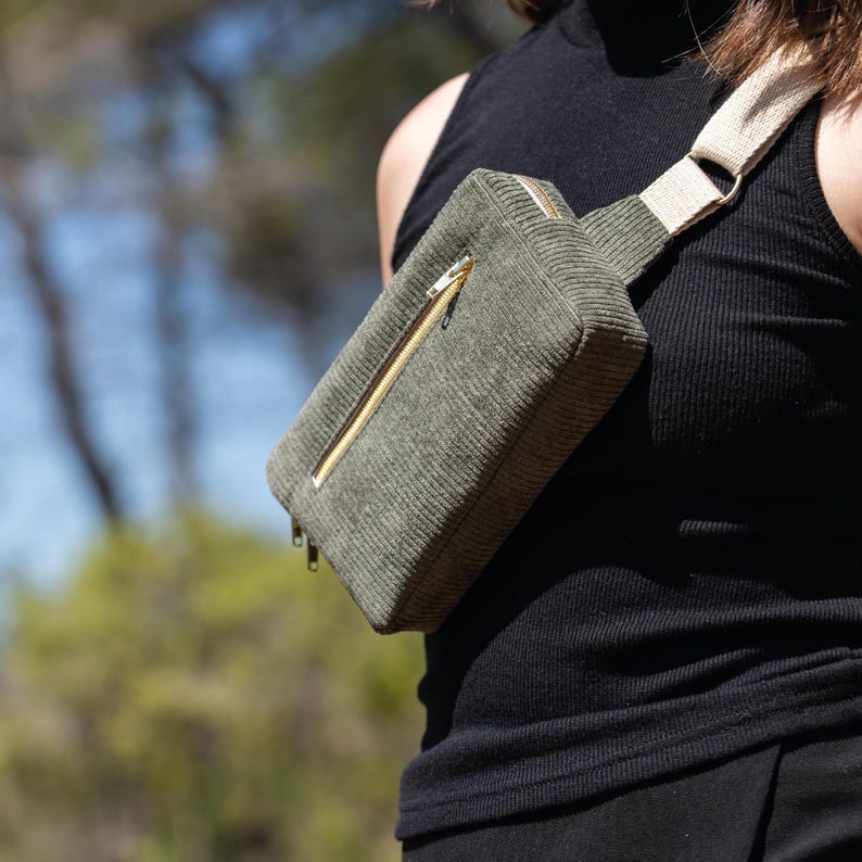 May include: Olive green corduroy fanny pack with a gold zipper. The bag has a beige strap and is worn across the body. The bag is rectangular and has a minimalist design. The fanny pack is a stylish accessory for carrying essentials.
