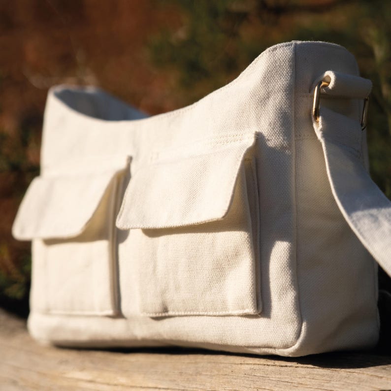 May include: A white canvas shoulder bag with two large front pockets. The bag has a long adjustable strap.