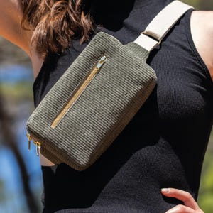 May include: Olive green corduroy fanny pack with a cream-colored strap and gold-toned zippers. The rectangular bag is worn across the body. The bag has a main compartment and a smaller front pocket.