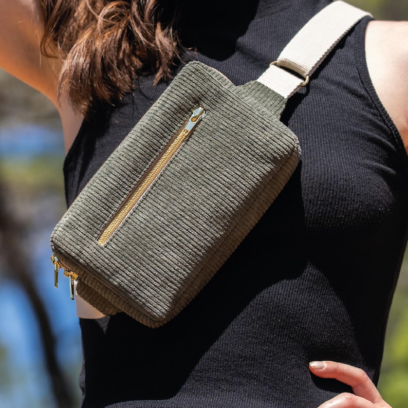 May include: Olive green corduroy fanny pack with a cream-colored strap and gold-toned zippers. The rectangular bag is worn across the body. The bag has a main compartment and a smaller front pocket.