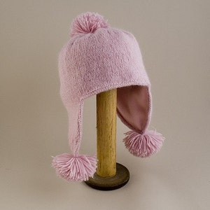 Puede incluir: Un gorro de punto rosa con orejeras y pompones. El gorro se muestra en un soporte de madera.