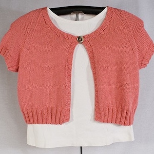 May include: A short-sleeved, coral pink knitted cardigan with a button closure. The cardigan is open in the front and is worn over a white t-shirt.