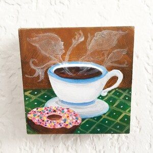 May include: A whimsical painting of a white teacup with a blue rim filled with dark coffee, steam rising above. A pink donut with colorful sprinkles sits in front of the cup. The background is brown with white ethereal shapes.