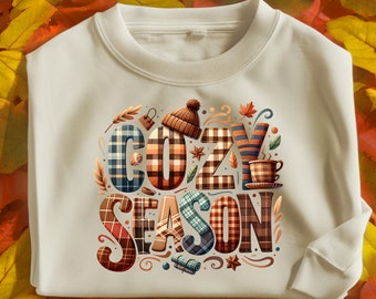 May include: A white sweatshirt with a graphic design of the words "Cozy Season" in a plaid pattern with fall-themed illustrations.