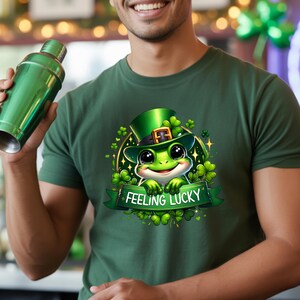 Pode incluir: Camiseta verde com um sapo de desenho animado usando um chapéu verde e cercado por trevos. O sapo segura uma faixa que diz "Feeling Lucky". A camiseta é de cor verde lisa.