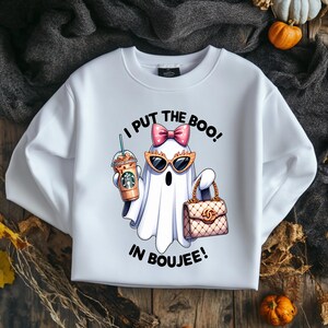 May include: A white crewneck sweatshirt featuring a graphic of a ghost wearing pink polka-dot bow and orange cat-eye sunglasses, holding a coffee cup and a quilted handbag. The text reads I PUT THE BOO IN BOUJEE in black capital letters.