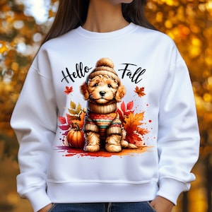 May include: White sweatshirt featuring a cute puppy wearing a knit hat and sweater, surrounded by fall leaves and a pumpkin. The words "Hello Fall" are written above the puppy in black script. The design has a watercolor effect.
