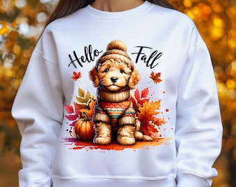 Może przedstawiać: Biała bluza z uroczym szczeniakiem w dzianinowej czapce i swetrze, otoczonym jesiennymi liśćmi i dynią. Słowa "Hello Fall" są napisane nad szczeniakiem czarnym pismem. Projekt ma efekt akwareli.